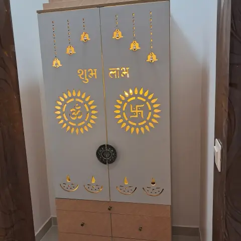 Freestanding mandir cabinet in a narrow alcove: grey shutters CNC-cut with backlit Om and swastik sunbursts, hanging bells
