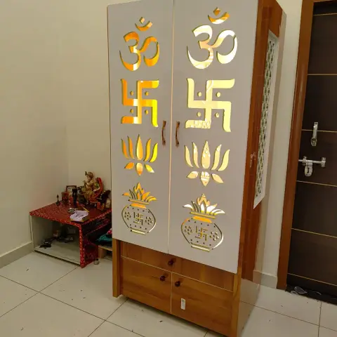 Freestanding pooja cabinet with mirrored gold-cut Om, swastik and lotus motifs on its doors, beside a small altar table with deity idols.