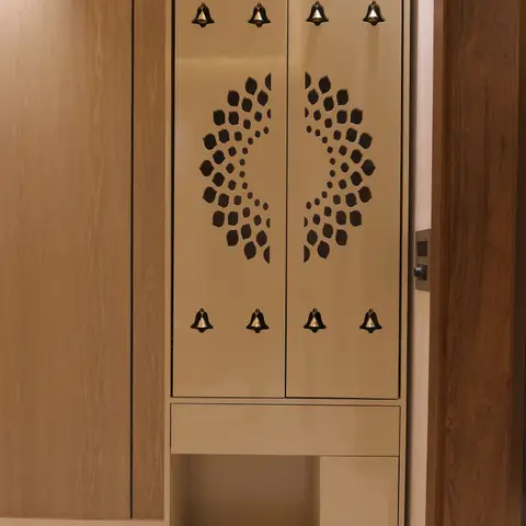 Freestanding pooja cabinet with taupe lacquered doors featuring a radial CNC-cut jaali pattern and hanging brass bells, with open shelving below.