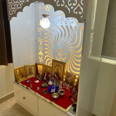 Freestanding pooja unit with a backlit jaali panel of an Om motif, deity photo frames, and brass ritual items on a red cloth