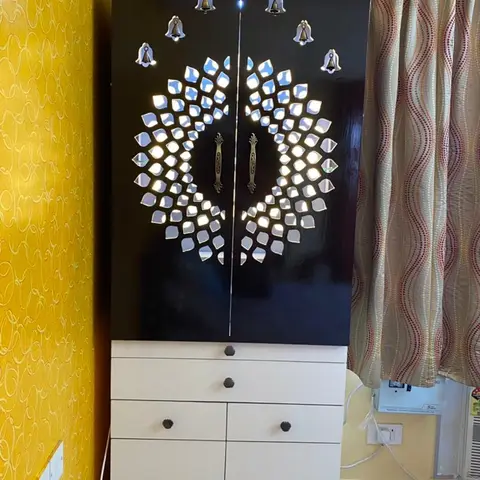 Freestanding pooja unit with a black lacquered peacock-motif backlit jaali door and hanging bell decorations, set against a yellow patterned wall