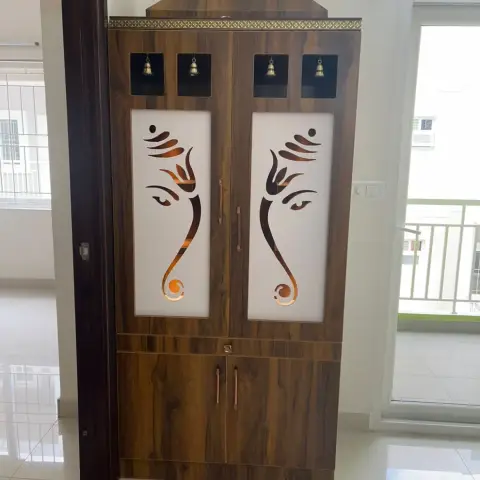 Freestanding walnut-laminate mandir unit with twin frosted shutters carrying CNC-cut Ganesha faces, four brass bells in cutout niches and a gold trim pelmet