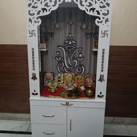 Freestanding white CNC-jaali pooja mandir with an engraved Ganesha backdrop and brass bells, set against a wallpapered dado wall.