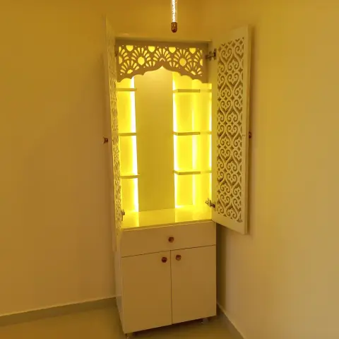 Freestanding white mandir with CNC jaali shutters swung open on a warm backlit niche of stepped shelves under a scalloped arch