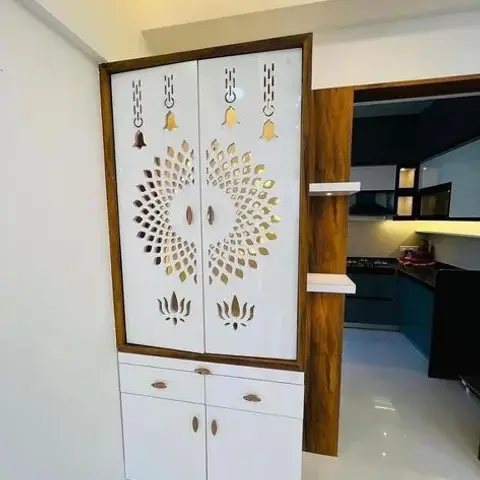 Freestanding white pooja cabinet framed in dark teak veneer, its upper shutters CNC-cut into a golden sunburst with lotus and hanging-bell motifs