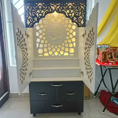 Freestanding white pooja cabinet with a backlit Om jaali panel, ornate perforated cabinet doors, and a dark drawer base