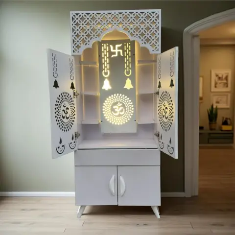 Freestanding white pooja mandir unit with cut-out jaali doors and a backlit Om and swastik motif panel, set against a sage green wall in a hallway nook.