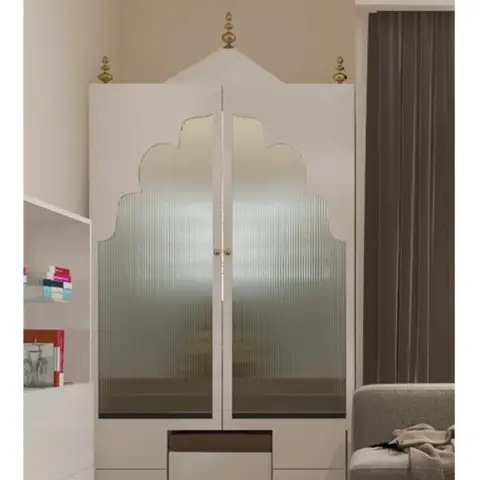 Freestanding white pooja unit with an arched mandir-shaped silhouette, fluted glass doors and brass finials, beside a living room sofa.