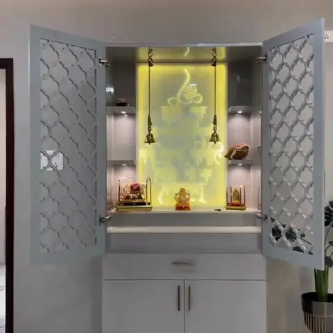Freestanding white pooja unit with jaali-patterned cabinet doors open to reveal a backlit yellow Ganesh silhouette panel, brass hanging bells