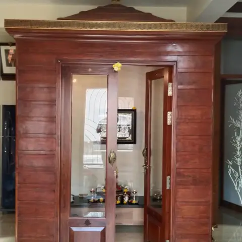 Freestanding wooden pooja unit with a gopuram-style pediment roof, glass-panelled doors, and a backlit diamond jaali panel above, deities visible inside.