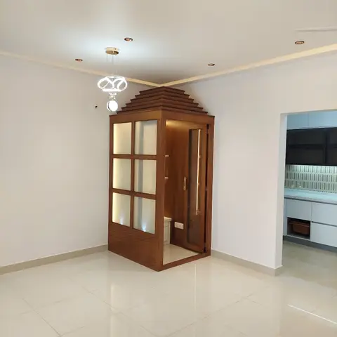 Freestanding wooden pooja unit with a tiered temple-style roof and frosted-glass shutters in the corner of an open-plan living area.