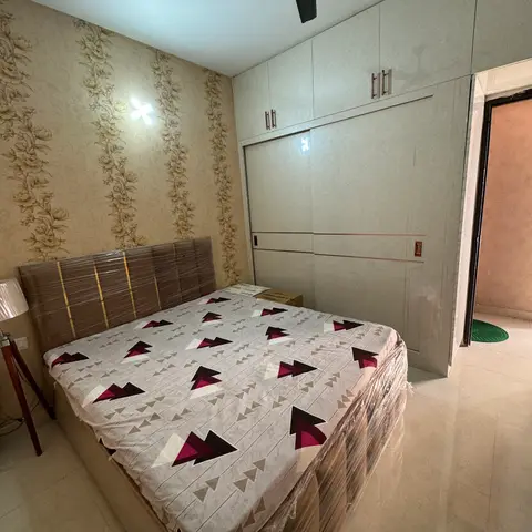 Freshly handed-over bedroom with the bed still shrink-wrapped in plastic, floral-stripe wallpaper behind the headboard and a full-height sliding wardrobe