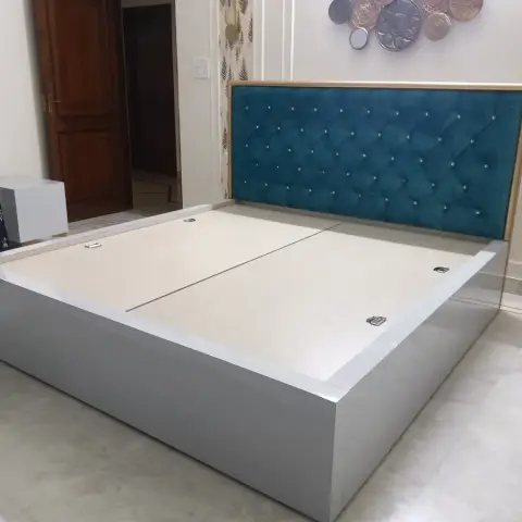 Freshly installed grey hydraulic storage bed with its lift-up deck still bare, backed by a teal velvet headboard tufted with crystal buttons inside a gold