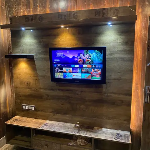 Freshly installed TV feature wall in dark wood-grain laminate with spotlit floating shelf and a drawer console below, still wrapped in protective film.