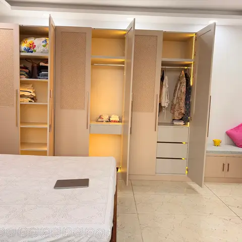 Full-height fitted wardrobe wall with internal shelf lighting, open shutters showing hanging rods and drawers, beside a cushioned window-seat nook.