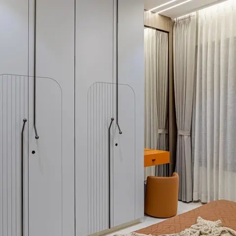 Full-height pale-grey wardrobe run with CNC-grooved arch detailing, next to a curtained dressing nook holding an orange laminate ledge and tan leather stool