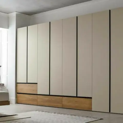 Full-wall hinged wardrobe in matt sand-beige with black shadow-gap reveals doing the work of handles and a band of oak drawer fronts at the base