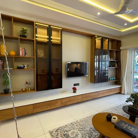 Full-wall wood-veneer TV unit with glazed display cabinets and an open bookshelf styled with plants and figurines, under a yellow LED cove-lit ceiling.