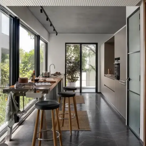 Galley kitchen running along a full-height garden glazing wall - a stainless-steel island with a timber worktop and three black-seat bar stools on jute