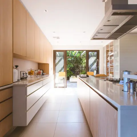 Galley kitchen with handleless light-oak cabinetry and a long stone island leading to glazed doors opening onto a deck