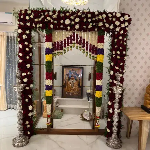 Glass-fronted home pooja niche dressed for a function — rose-and-marigold garlands framing the teak-and-glass doors