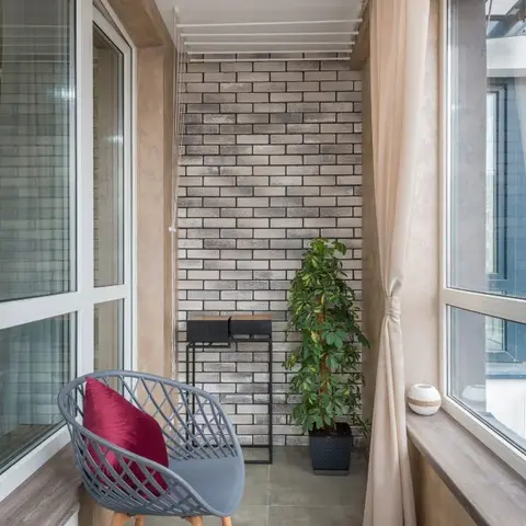 Glazed balcony with a brick-effect accent wall, a woven grey accent chair with a red cushion, a small side table and a potted plant.