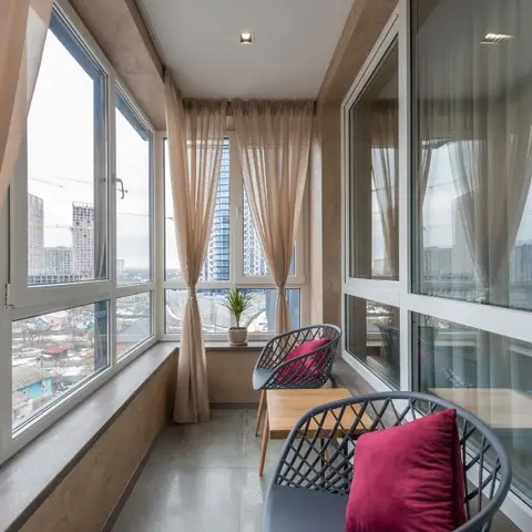 Glazed high-rise balcony with sheer beige curtains, a small wood side table, and grey woven-frame chairs with maroon cushions overlooking the city skyline.