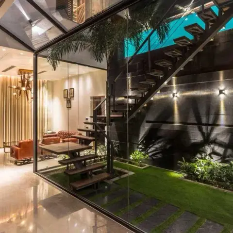 Glazed internal courtyard at night: cantilevered timber treads climbing a dark stone-clad wall over a grass strip with inset stone pavers and uplit palms