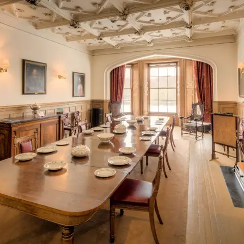 Grand panelled dining hall with an ornate plaster ceiling, portrait-hung walls and a long antique table set with china.