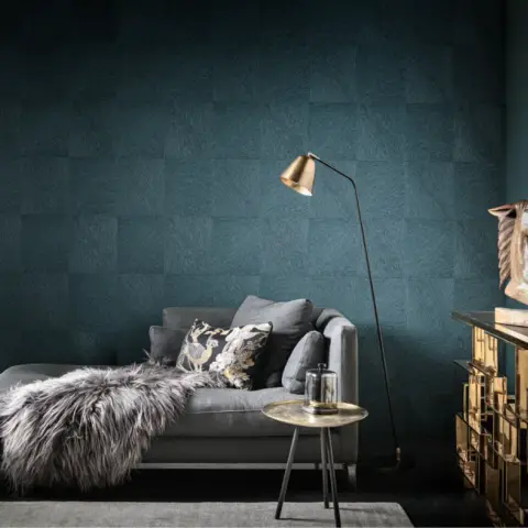 Grey chaise sofa with a shaggy sheepskin throw set against a full-height teal wallpaper laid in feathered square blocks, with a brass arc floor lamp