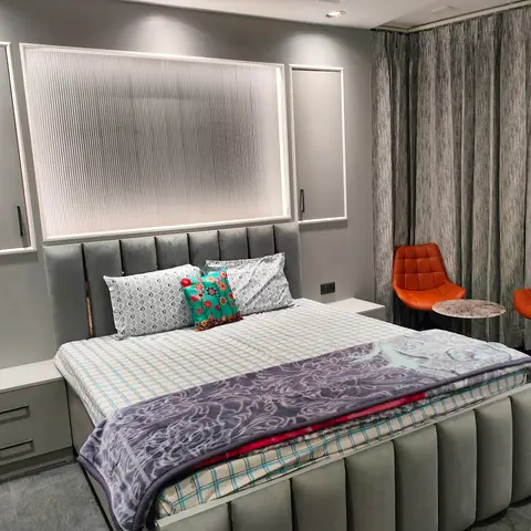 Grey channel-tufted bed against a backlit fluted-glass headboard panel, with a small orange-chair seating nook by the curtained window.