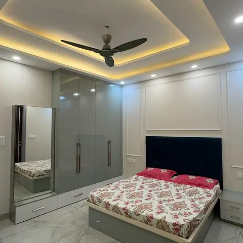Grey glossy wardrobe with full-length mirror beside a navy tufted-headboard bed under an arrow-shaped cove-lit false ceiling, marble floor