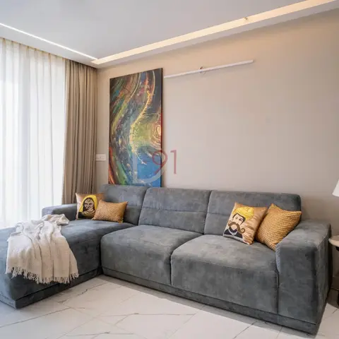 Grey L-shaped sectional sofa in a living room with a cove-lit ceiling, marble-look tile flooring, and gold turban-motif cushions against a beige accent wall.