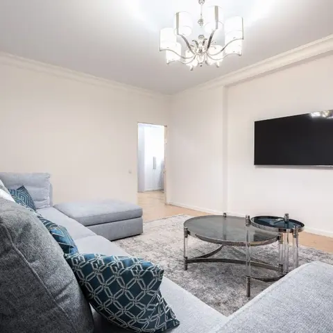 Grey L-shaped sectional sofa with teal patterned cushions facing a wall-mounted TV, under a chrome drum chandelier and glass coffee table.