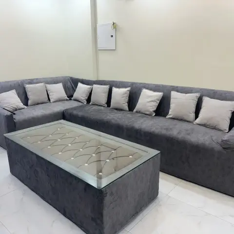 Grey L-shaped sectional sofa with nine matching cushions, paired with a tufted glass-top coffee table in an otherwise bare living room.