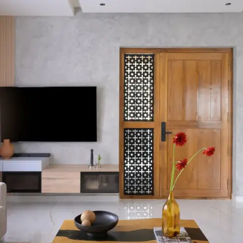 Grey lime-plaster TV wall with an oak fluted return, faced across the sofa by a solid teak main door paired with a black square-lattice metal screen and the