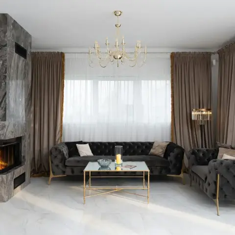 Grey marble-clad corner fireplace with a live fire beside a charcoal velvet button-tufted sofa and matching loveseat on white marble floor
