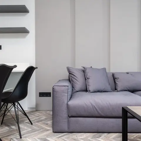 Grey-panelled living room corner with an L-shaped grey sectional sofa, herringbone wood flooring, and black Eames-style chairs at a small desk nook