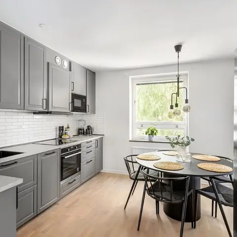 Grey shaker-style kitchen with white subway-tile backsplash and black fixtures, opening to a round black dining table with woven chairs by a window.