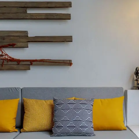 Grey sofa corner styled with mustard and graphic-print cushions beneath a rustic reclaimed-wood wall art piece and a vintage sewing machine on a plinth.