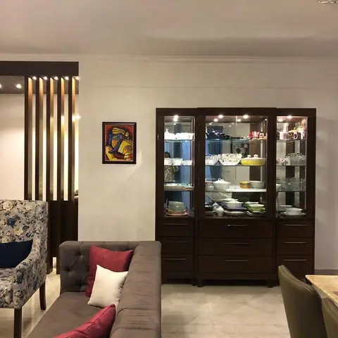Grey tufted sofa with maroon cushions facing a tall lit wood-and-glass crockery display cabinet, beside a fluted wood partition screen.
