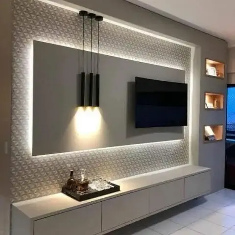 Grey TV wall framed by a backlit 3D-triangular panel border, with three slim black cylinder pendants hanging beside the screen over a floating white drawer