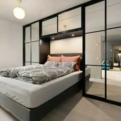 Grey upholstered divan bed tucked under a dark bridge unit, framed by black-gridded mirrored sliding wardrobe doors that reflect the corridor beyond.