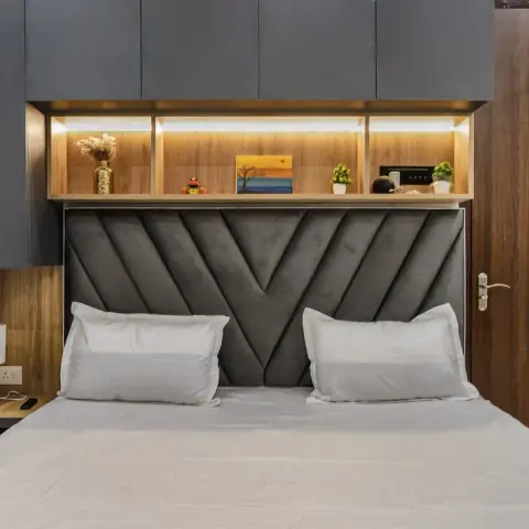 Grey V-channelled upholstered headboard under a backlit wood-laminate display niche, with graphite loft shutters above and a lamp on the built-in side table