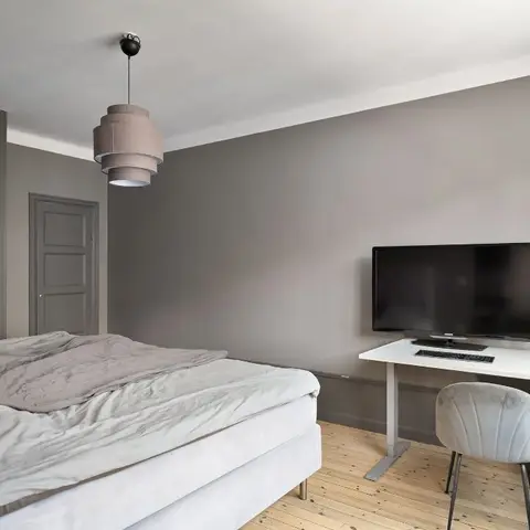 Grey-walled bedroom with a flush handleless wardrobe wall, a tiered dusty-pink drum pendant and a white sit-stand desk with two monitors on raw pine boards.