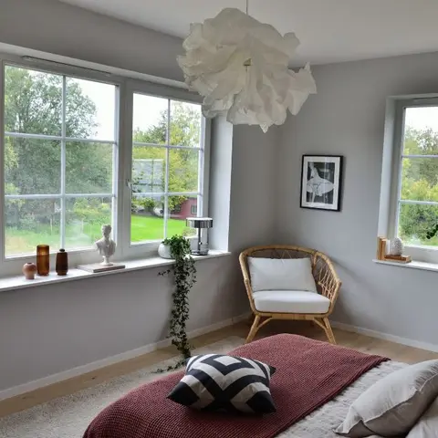 Grey-walled corner bedroom where a rattan armchair with a white cushion sits between two windows lined with vases, a bust and trailing ivy