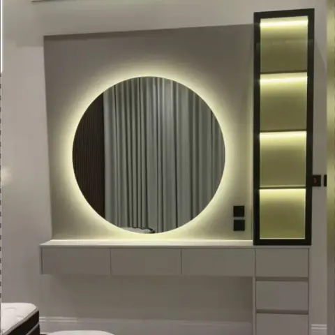 Halo-lit round mirror over a wall-hung greige dressing counter with a drawer bank, next to a black-framed column of four warm-lit display niches