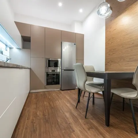 Handleless taupe kitchen with an integrated oven and fridge tower opening into a two-seat dining nook against a wood-panelled accent wall.