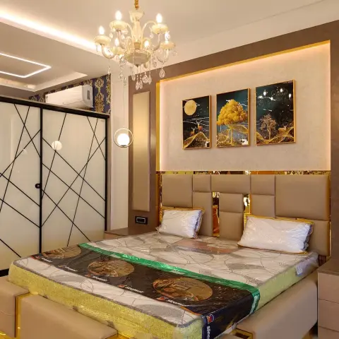 Handover-day bedroom with the mattress still in factory wrap: beige upholstered headboard panels edged in gold mirror below a backlit triptych of gold-tree