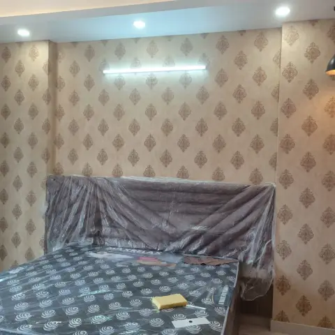 Handover-stage bedroom with the bed and its tall headboard still shrink-wrapped in plastic, against a beige damask wallpaper